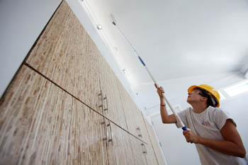 Photo of a young woman using an paintbrush extension to paint the ceiling of a room.