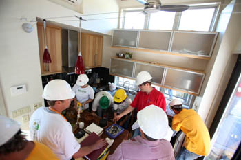 Photo of a group of people standing in a kitchen. A man in a red shirt points to something beneath a counter as the others look on.