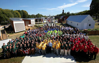 Photo of a large group of people standing together in the solar village. Subgroups wear different colors of matching shirts to identify them as a team. In the front is Solar Decathlon Director Richard King.