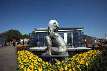 Photo of a statue of a person holding a sphere and surrounded by flowers. In the background is the Team Spain house and visitors waiting to tour it.