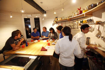 Photo of two people cooking in a kitchen with a round wall. Around a table in the center, six students converse.