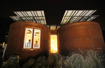 Photo of the exterior of the Cornell house at night. Through a lighted window, two people are visible.
