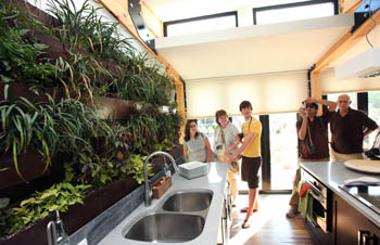 Photo of several people standing at the end of a galley kitchen. On the left wall, above the sink, is a variety of live plants.
