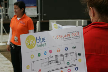 Photo of a woman holding a sign that says "s.kyblue" and includes a phone number for an audio tour. A member of the Solar Decathlon visitor liaison staff is in the background.
