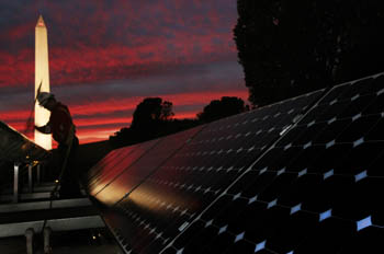 Photo of a person cleaning a long line of PV panels on the roof of a house. In the background is the illuminated Washington Monument and a pink sunset.