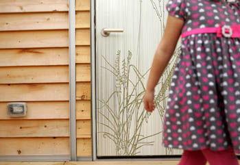 Photo of the body of a child as she walks by a door of a wood-sided house. On the door is the image of tall grass.