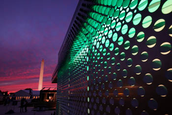 Photo of one side of the Virginia Tech house, which includes a sliding siding of punched metal. Green light illuminates through the holes in the siding. The Washington Monument is seen in the background.