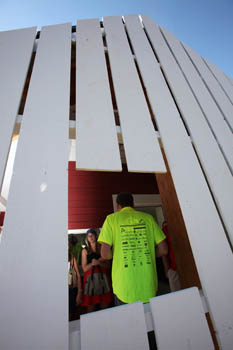 Photo of an exterior wall of a house, which is composed of vertical wood boards. Through a window cut in the boards, a student can be seen giving a tour to visitors.