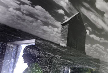 Photo of a black-white-picture of a barn in a field. Reflected in the glass is the silhouette of a young man standing near a bright doorway.