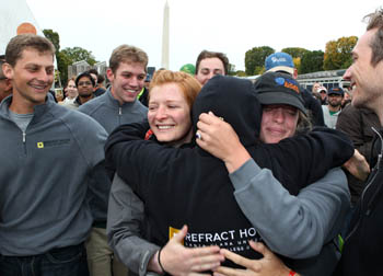 Photo of two young woman hugging a man, whose back is to the camera. A group of young men around them smile.