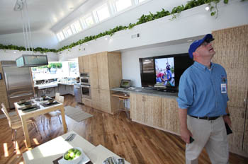 Photo of a man walking through the living area of a house and looking up. A television, kitchen, and dining area are visible in the house.