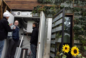 Photo of three men standing within a set of open doors that expose the mechanical room of the Penn State House. Surrounding the doors are live plants integrated into the exterior siding of the house.