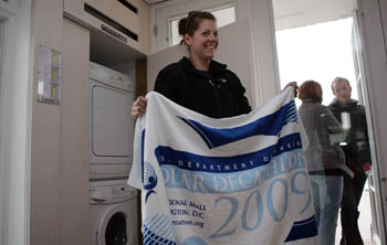 Photo of a young woman standing near a clothes washer and dryer and holding a towel with Solar Decathlon branding.