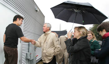 Photo of two men shaking hands while a woman holding an umbrella looks on.
