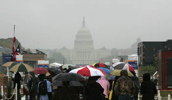 Photo of people carrying umbrellas crowding Decathlete Way in the solar village. The U.S. Capitol is seen through foggy skies in the background.