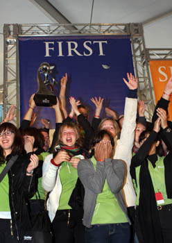 Photo of a group of students celebrating in front of a first-place award banner. The first-place trophy is lifted above their heads, and most students extend their arms in the air.