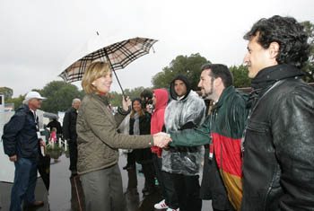 Photo of Princess Cristina shaking hands with a line of students. She is holding an umbrella to shield herself from rain.