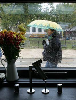 Photo looking outside from the inside of the University of Wisconsin-Milwaukee's house. Inside, a faucet and urn of plants are visible on a counter. Outside, a woman walks by carrying an umbrella.