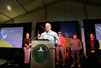 Photo of Richard King speaking from behind a lectern with the Department of Energy seal. Behind him stand five people: the Solar Decathlon competition manager and four people who represent the students of each Solar Decathlon.