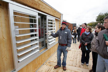 Photo of a young man standing on a deck next to louvered windows. He is speaking to a group of visitors.