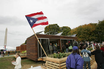 Photo of the Puerto Rico house, which has a Puerto Rican flag flying outside. A line of people waits to tour the house.