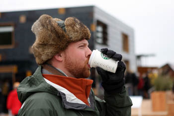 Photo of a man in a fur-trimmed hat who is bundled up and drinking a coffee. The Team Germany house is visible in the background.