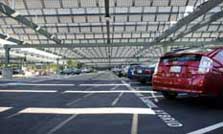 Photo of a metal structure over a paved parking lot with cars. The structure supports photovoltaic panels and shades the cars beneath. The nearest parking spaces are labeled "Carpool Hybrid."