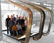 Photo of 16 members of the Arizona team standing inside the metal skeleton of their house. The skeleton and the team members are in color. The background is black and white. In the background are the walls of their construction area and buildings, which can be seen through windows.