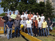Photo of 18 people wearing jeans and various types of t-shirts. They are standing in the sun between two widely- spaced wooden floor joists on the ground. Blue sky and trees are in the background.