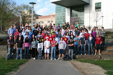 Photo of more than 100 casually dressed college students standing as a group in front of a large building with a glass front.