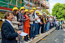 Photo of approximately 25 members of Team Germany wearing yellow hard hats and standing side-by-side in front of construction scaffolding.