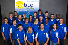 Photo of 21 members of the Kentucky team, all of whom are wearing blue collared shirts with the University of Kentucky logo on the left chest. Behind the team is a promotional sign for the s•ky blue house.
