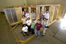 Photo of 11 members of the University of Minnesota team standing in front of and inside the wooden frame of their team house. The house is inside a large warehouse-like building.