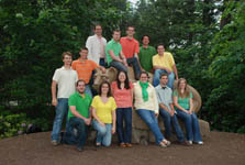 Photo of thirteen people—each of whom is wearing an orange, green, yellow, or white shirt—sitting on, standing near, and kneeling in front of a large brown statue of a lion. Green trees and foliage are in the background.