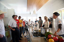 Photo of a group of visitors inside Team Austin's kitchen, which is filled with sunshine that reflects against a gleaming wooden ceiling panel. Lights hang down above a wooden island that contains a stovetop, pots and pans, and a basket of fresh fruit.