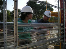 Photo of two students wearing hard hats, who are installing long glass tubes in vertical rows along metal fittings on the outside of their team's house.