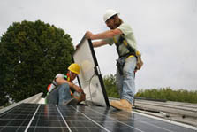 Photo of two students in hard hats installing solar panels on their house's roof. One student holds the large solar panel while the other adjusts the wiring in the back of the panel. The panels already installed face up to the sun and are glossy black with white grid marks.