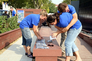 Three  Penn State students (wearing matching blue t-shirts) lay brochures about their team's home on a bench.  One student gives another a "bear hug" when they learn they won third-place in the 2007 Solar Decathlon Communication contest.