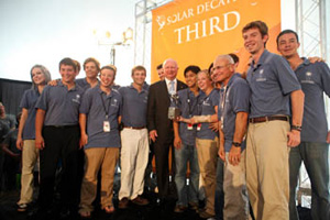 Photo of 15 people in team uniforms standing around a man in a suit in front of a third-place banner.
