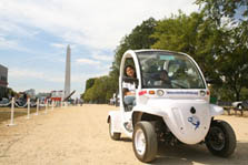 Photo of two smiling female students inside a white electric car. The Washington Monument looms in the background.