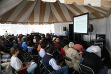 A man presents information about energy-efficient buildings to a group of people seated under a canopy at the 2007 Solar Decathlon.