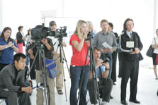 Photo of a group of people standing in a bright white tent. Some are operating video cameras or holding microphones. Others are taking notes in small notebooks.