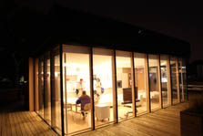 Photo of four people inside a brightly lit house with glass walls on three sides. Outside, it is dark.