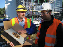Photo of two men wearing hard hats and safety vests looking at a metal meter box within a cardboard box.